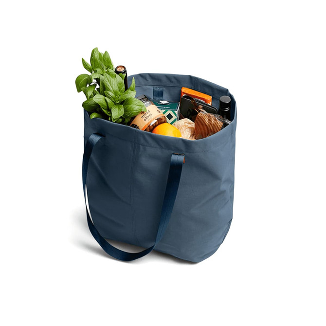 A navy blue tote bag filled with fresh basil, groceries, and a drink bottle. The bag has sturdy handles and a logo.