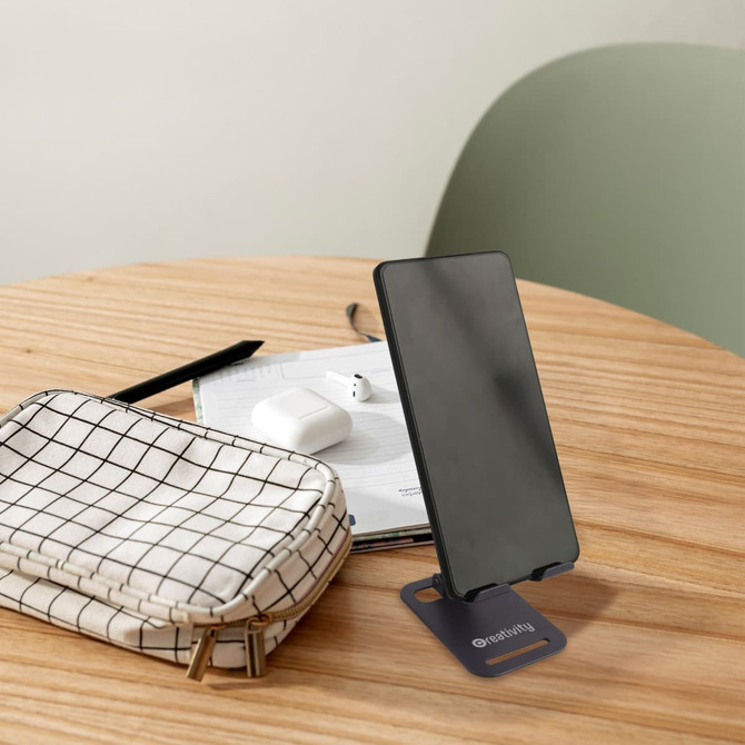 A foldable metal mobile phone stand in black, with a smartphone displayed, on a wooden table along with a bag and accessories.