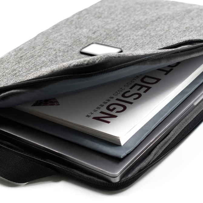 A grey laptop sling bag with a zip, containing a laptop and a design book. It features a small logo.