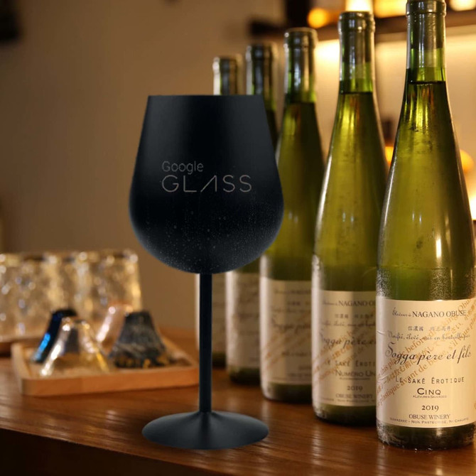 A black stainless steel wine glass beside several green wine bottles on a wooden surface. The glass has a branding logo.