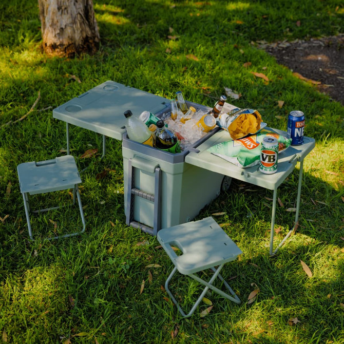 A grey cooler box sits on grass, featuring a fold-out table and two small chairs, filled with drinks and snacks.