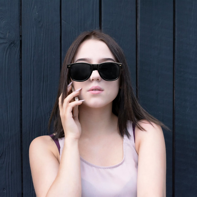A pair of black classic sunglasses displayed against a dark wood background, worn by a young woman.