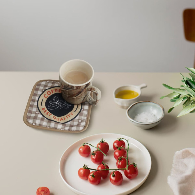 A medium quilted pot holder in a checkered pattern with a logo, placed on a table with a mug and fresh tomatoes.