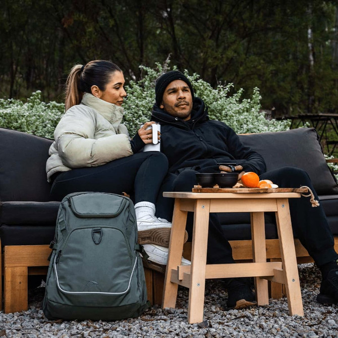 A green High Sierra Access 4.0 backpack sits on the ground beside a couple relaxing on a couch outdoors.