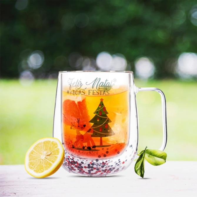 A Christmas-themed double wall mug with a tree and reindeer design, filled with a drink and a lemon slice.