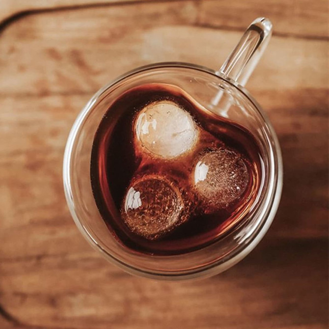 A heart-shaped double wall mug filled with dark liquid and three ice cubes, set on a wooden surface.