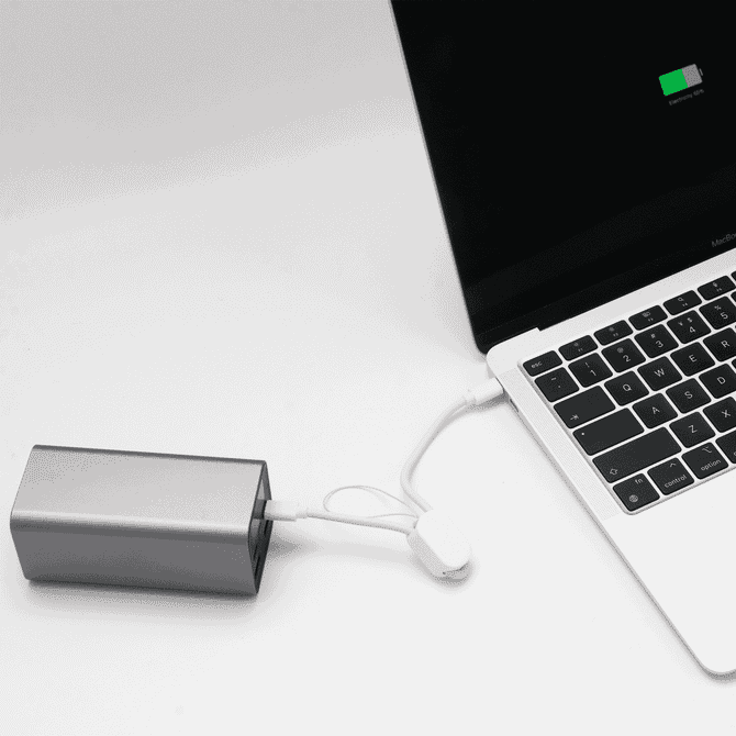 A silver power bank connected to a laptop via a white charging cable. The laptop screen shows a battery charge indicator.