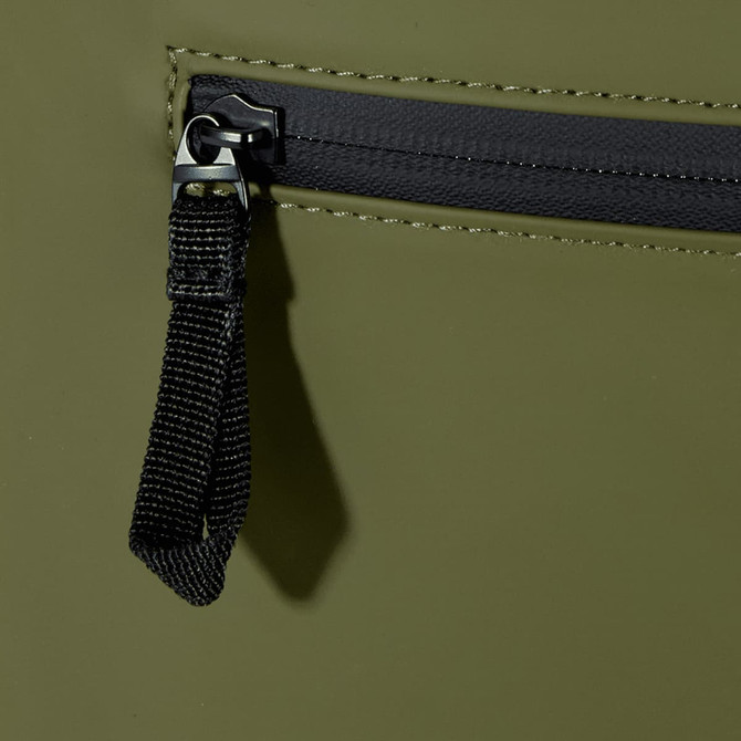 A close-up of a dark olive green roll-top backpack featuring a zippered pocket and a black zipper pull.