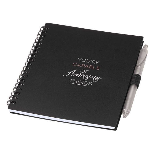 A black spiral-bound journal with a silver pen attached. The journal features motivational text and has a logo.