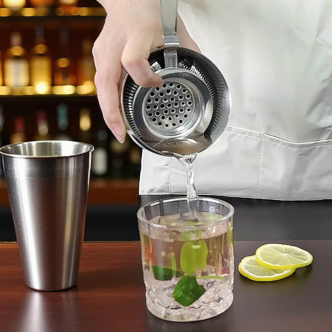 A round silver cocktail strainer pours a drink into a glass with ice and mint, alongside lemon slices.