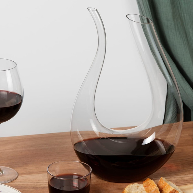 A U-shaped glass wine decanter with red wine, alongside wine glasses and bread on a wooden table.