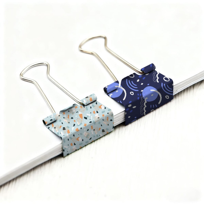 Two decorative binder clips in light blue and dark blue patterns sitting on a stack of white paper.