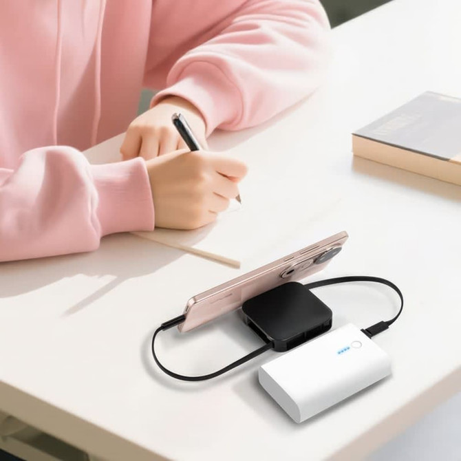 A white retractable fast charge cable and stand connected to a smartphone on a desk, with a person writing in the background.