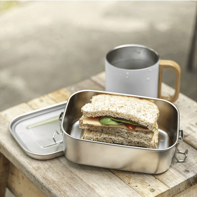 A stainless steel lunch box with a sandwich inside, placed beside a drink bottle on a wooden surface.