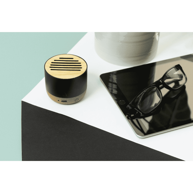 A wireless speaker in natural wood and black, positioned on a white surface next to a tablet and glasses.