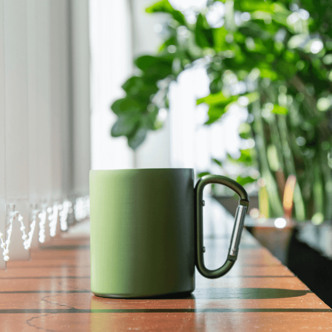 A green thermo mug with a carabiner handle, placed on a wooden surface with plants in the background.