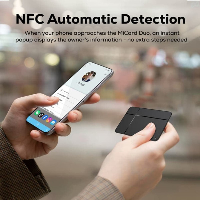 A person holds a black tracking tag near a smartphone displaying contact information. The background is blurred.
