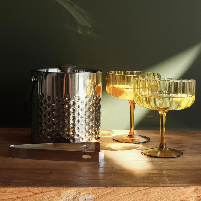 A stainless steel ice bucket with a textured design, accompanied by two amber cocktail glasses and a silver ice tongs.