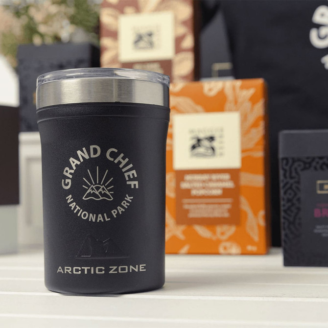 A black insulated drink tumbler with a silver lid, surrounded by colourful packaging. Includes a branded logo.