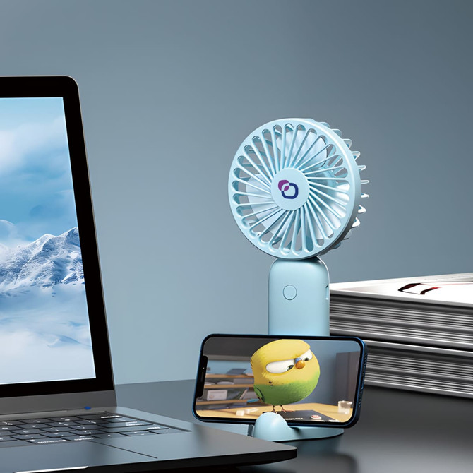 A light blue handheld fan with a phone stand, positioned next to a laptop and stacks of magazines.