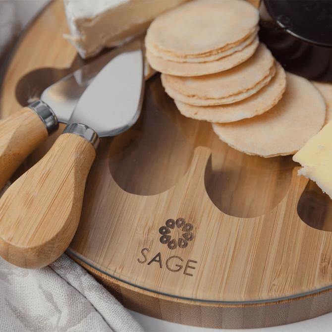 A bamboo cheese board featuring round crackers, cheese, and serving tools. Includes a logo on the board.