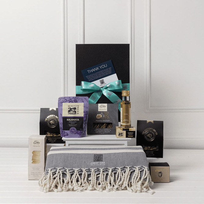 A gourmet gift pack featuring assorted food items, a stylish grey throw, and a bottle, all presented in a black box.