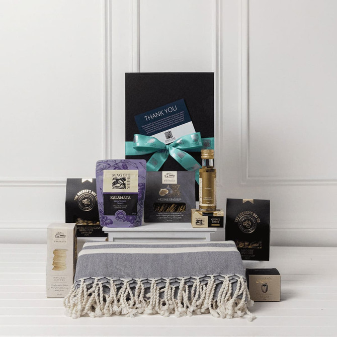 A gourmet gift pack featuring a grey and white throw, assorted treats, and a small bottle, all in elegant packaging.