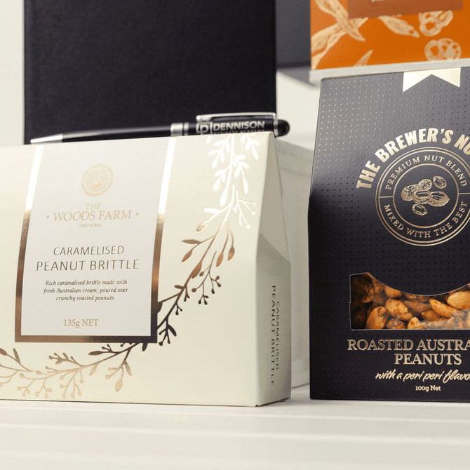 A white box of caramelised peanut brittle and a black bag of roasted Australian peanuts, with a pen beside them.