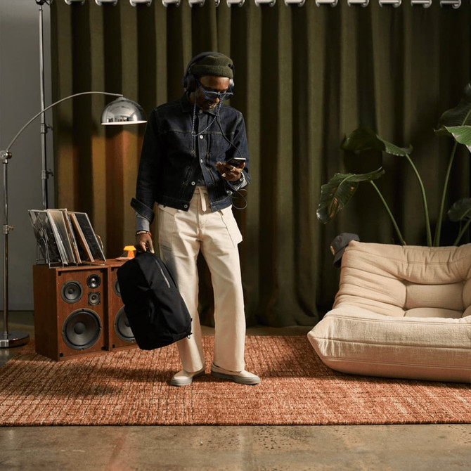 A person in casual attire holds a black backpack while standing in a room with speakers, plants, and a rug.