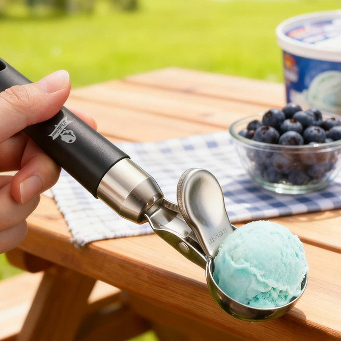 A black ice cream scoop with an easy trigger holds a scoop of blue ice cream, beside blueberries and a tub of ice cream.