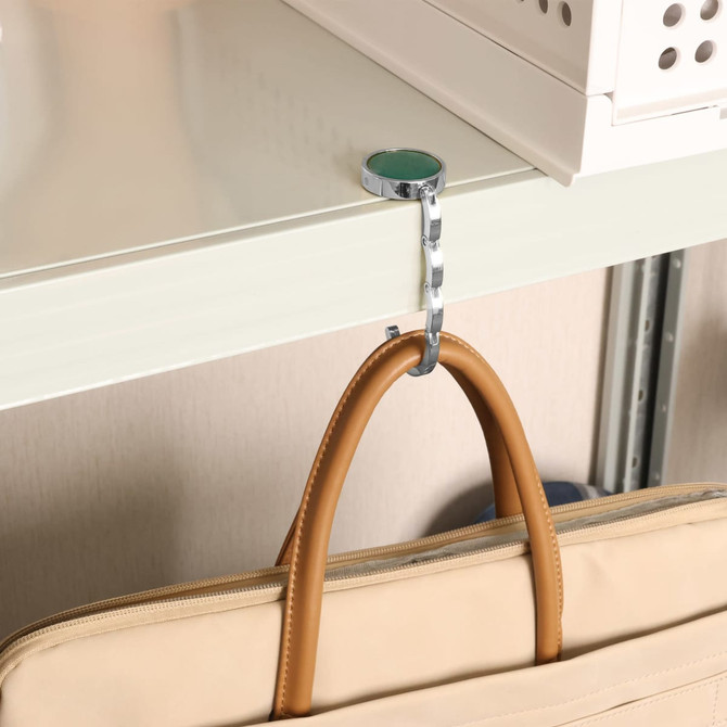 A round foldable desk hook in silver, attached to a shelf, with a tan handbag hanging from it.