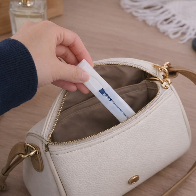 A hand placing a tube into a cream-coloured handbag on a wooden surface. The bag has gold hardware.