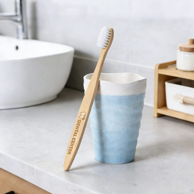 A large bamboo toothbrush with a light wooden handle and white bristles rests against a blue ceramic cup on a countertop.