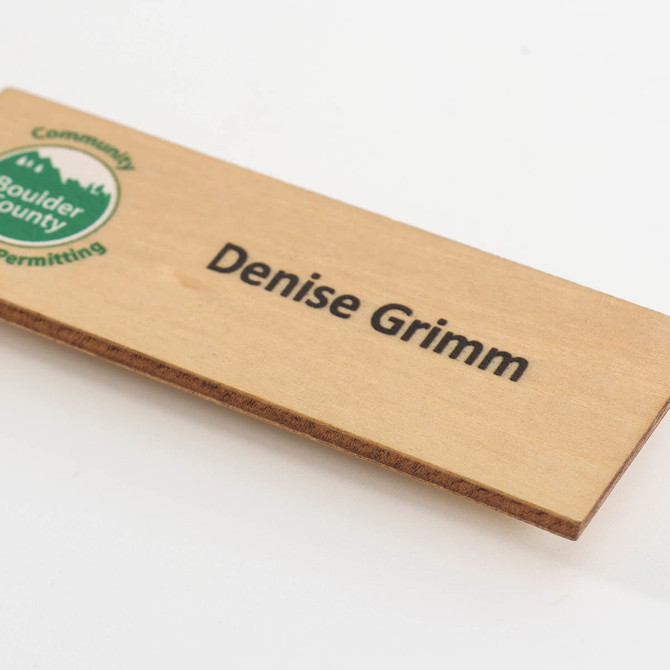 A wooden name badge in a natural finish featuring a logo and black text.