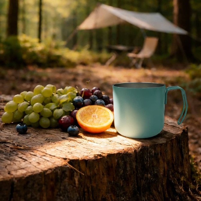 A stainless steel insulated mug in teal sits beside a mix of grapes and an orange on a wooden stump in a forest setting.