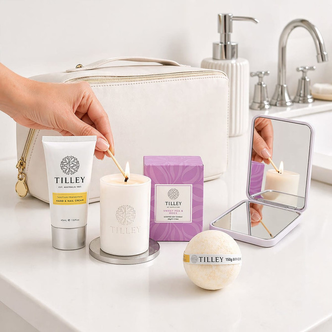 A cream toiletry bag with a candle, bath bomb, hand cream, and a compact mirror, arranged on a bathroom countertop.