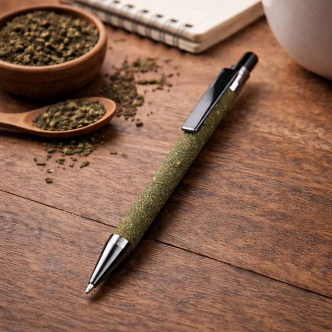 A green pen with a silver tip rests on a wooden surface, surrounded by tea grounds and a notebook.