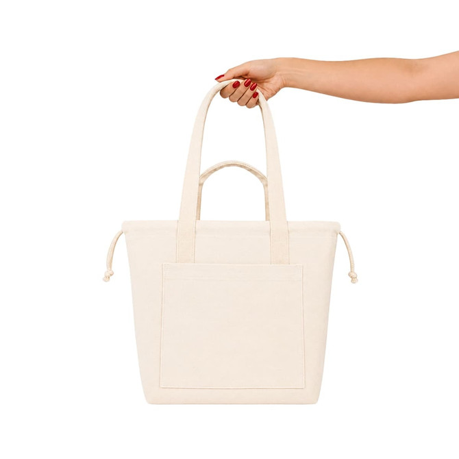 A cream tote bag with two handles and a front pocket, held by a hand with red nails. It has a logo.