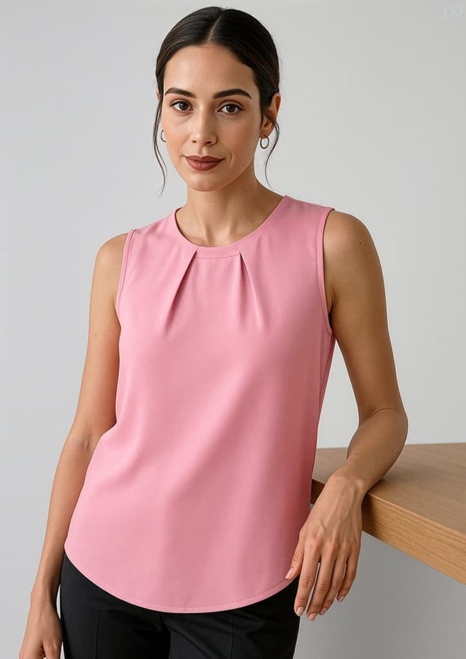 A pink sleeveless blouse with pleat detail, styled with a model standing beside a wooden surface.