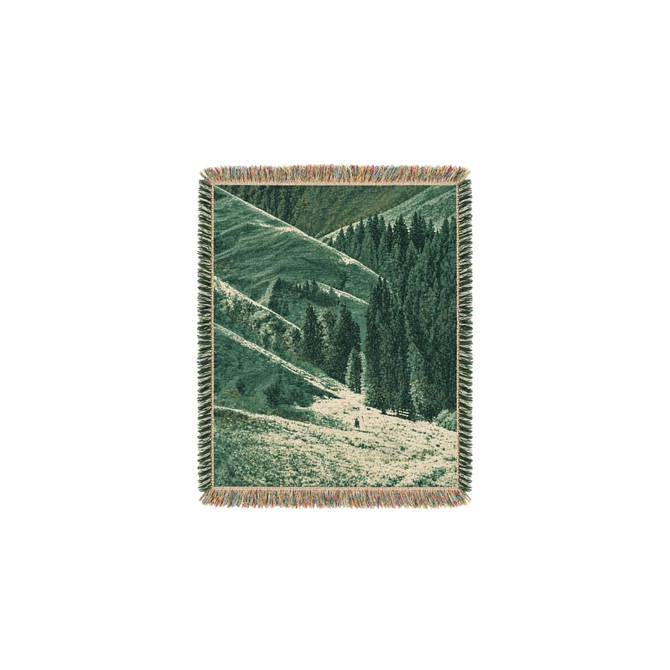 A tapestry blanket featuring green and earthy tones depicting rolling hills and tall trees. Fringed edges complete the design.