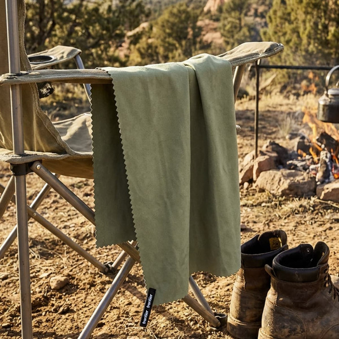 A green Frontier Rapid Dry Towel hangs from a camping chair beside a fire, with boots on the ground nearby.
