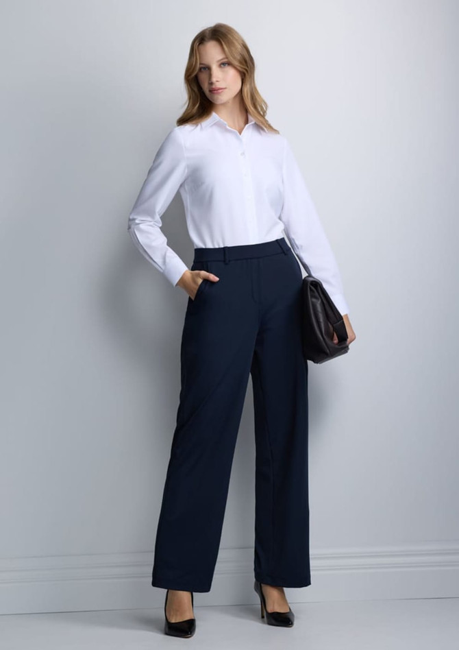 A woman wears a white long-sleeve shirt and navy trousers, holding a bag, posing against a plain background.