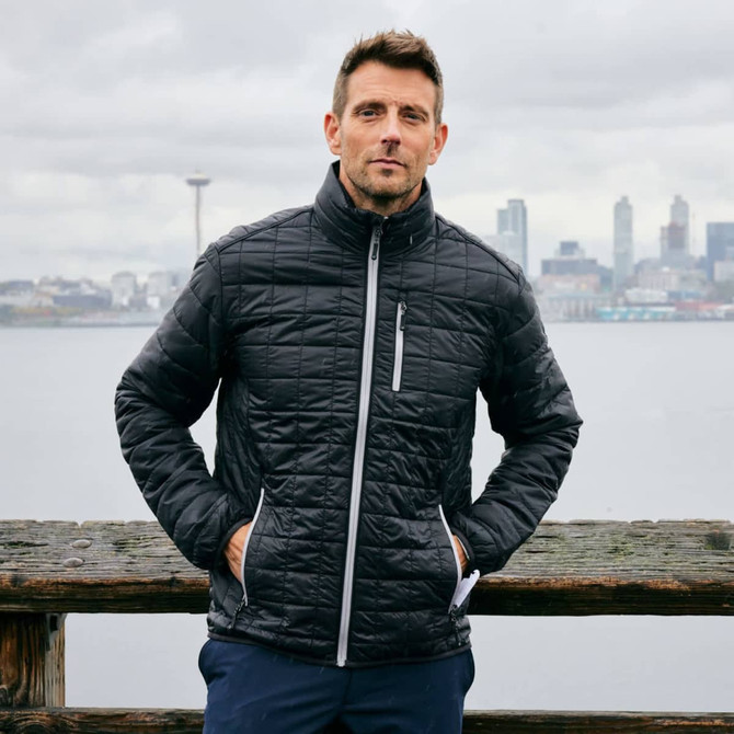 A man stands by a waterfront wearing a black Cutter & Buck Rainier men's jacket with a zippered pocket.