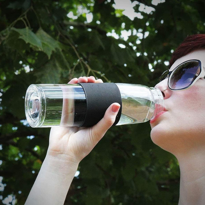 A drink bottle with a clear body and a black silicone grip, held by a person sipping water outdoors.