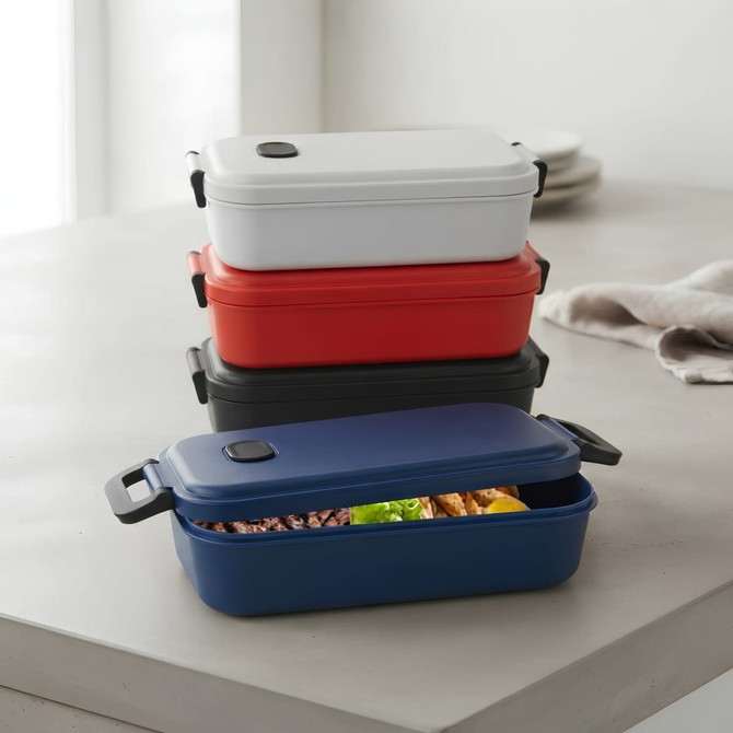 A stack of four lunch boxes in red, white, navy, and blue, featuring a lid and a small logo on each.