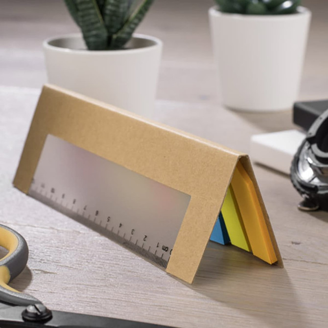 A ruler-shaped sticky note pad in yellow, blue, and green, presented in a brown cardboard holder with a logo.