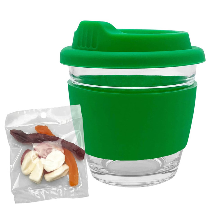 A green glass coffee cup with a silicone lid and sleeve, accompanied by a packet of mixed lollies in various colours.