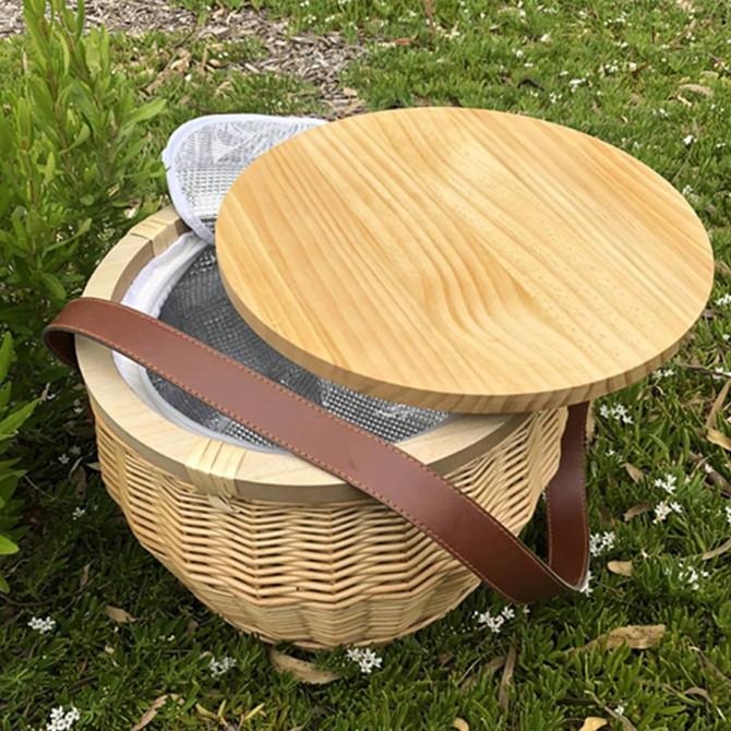 A round wicker picnic cooler basket in natural tones, featuring a wooden lid and a brown leather strap.