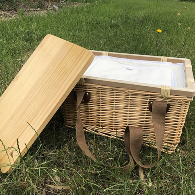 A square wicker picnic cooler basket in natural tones with a bamboo lid and brown straps, placed on grass.