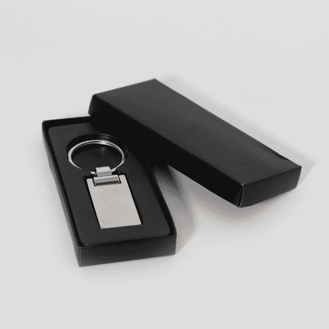 A silver keychain displayed inside a black box. The keychain has a rectangular shape with a polished finish.